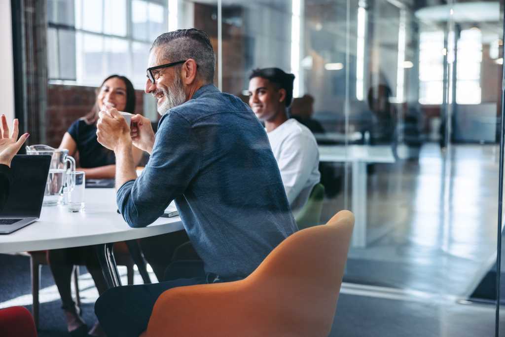 Happy mature businessman attending a meeting with his colleagues in an office. Experienced businessman smiling cheerfully while sitting with his team in meeting room. Businesspeople working together